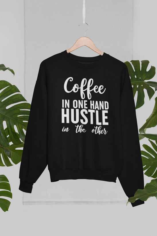 Coffee In One Hand Hustle In The Other Sweat Shirt displayed on a hanger with green plants in the background.