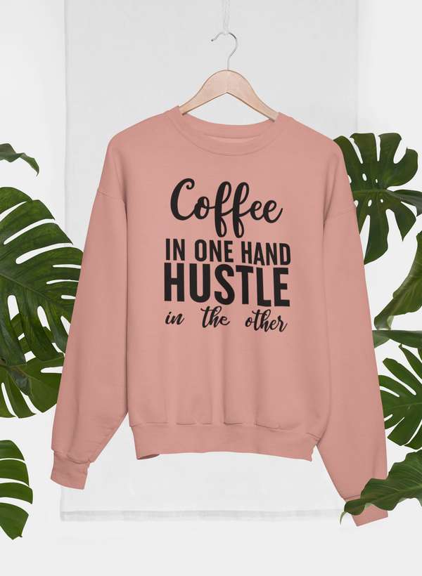 Coffee In One Hand Hustle In The Other Sweat Shirt hanging on a rack with green leaves in the background.