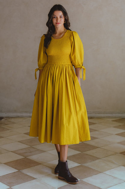 Freya Smocked Linen Midi Dress in yellow with puff sleeves and smocked bodice, set in a stylish indoor setting.